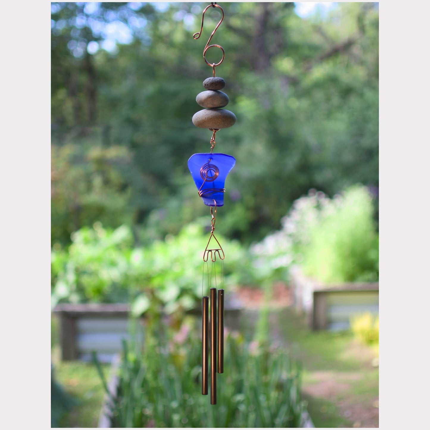 Decorative wind chime with blue glass piece and pebble design against a garden background