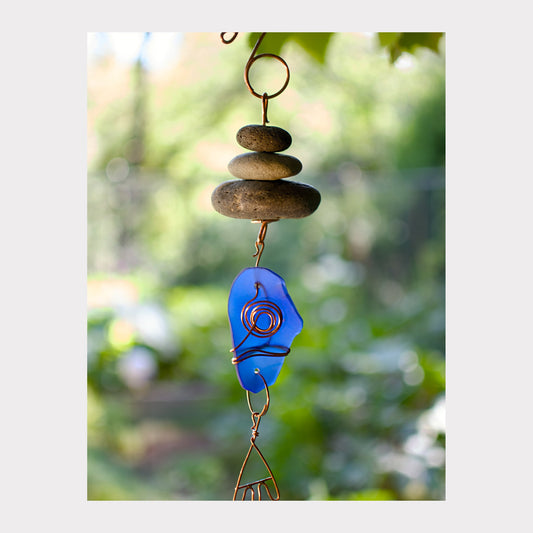 Decorative hanging with blue glass piece and stones against a blurred green background